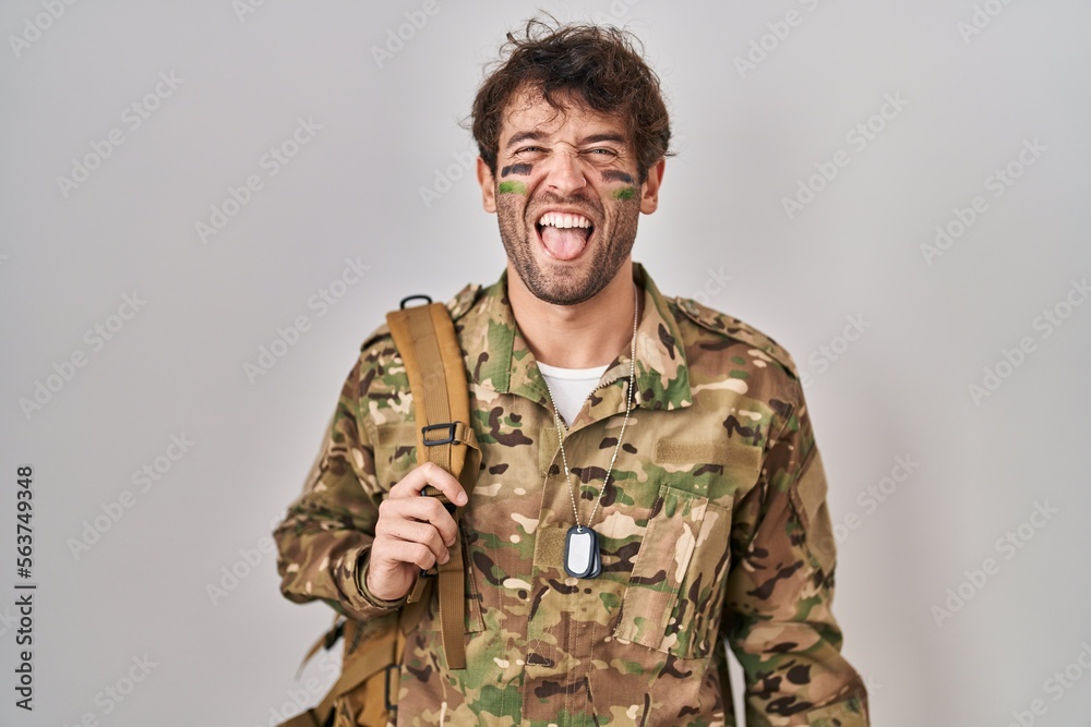 Hispanic young man wearing camouflage army uniform sticking tongue out happy with funny expression. emotion concept.