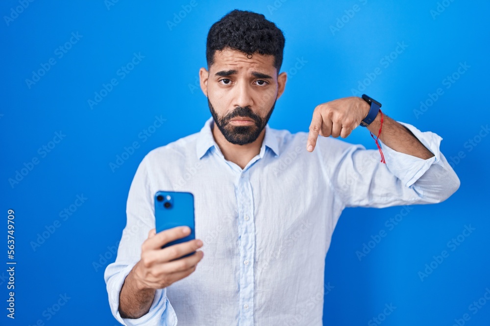 Hispanic man with beard using smartphone typing message pointing down looking sad and upset, indicating direction with fingers, unhappy and depressed. Hispanic man with beard using smartphone typing message pointing down looking sad and upset, indicating direction with fingers, unhappy and depressed.
