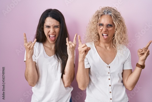 Mother and daughter standing together over pink background shouting with crazy expression doing rock symbol with hands up. music star. heavy music concept.