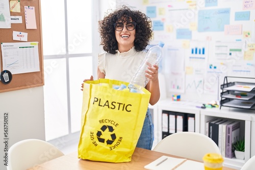 Young middle east woman holding recycling bag with plastic bottles at the office smiling and laughing hard out loud because funny crazy joke.