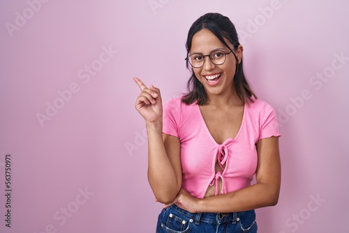Hispanic young woman standing over pink background wearing glasses with a big smile on face, pointing with hand and finger to the side looking at the camera. © Krakenimages.com