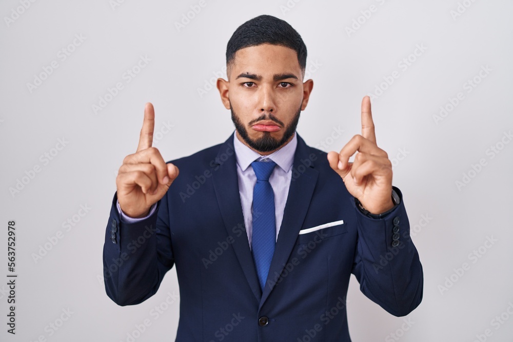 Young hispanic man wearing business suit and tie pointing up looking sad and upset, indicating direction with fingers, unhappy and depressed. Young hispanic man wearing business suit and tie pointing up looking sad and upset, indicating direction with fingers, unhappy and depressed.