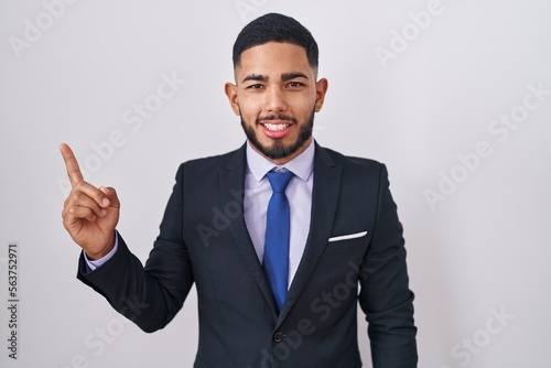 Young hispanic man wearing business suit and tie with a big smile on face, pointing with hand finger to the side looking at the camera.