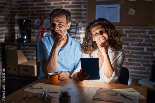 Middle age hispanic couple using touchpad sitting on the table at night looking confident at the camera smiling with crossed arms and hand raised on chin. thinking positive.