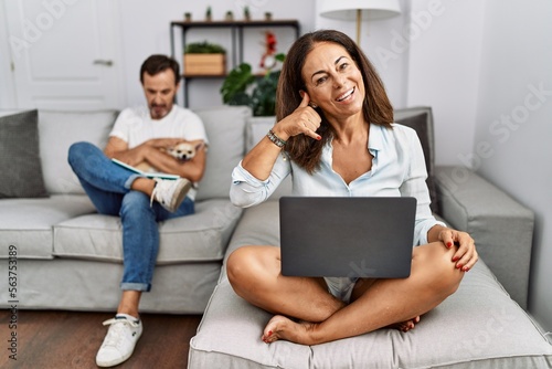 Hispanic middle age couple at home, woman using laptop smiling doing phone gesture with hand and fingers like talking on the telephone. communicating concepts.