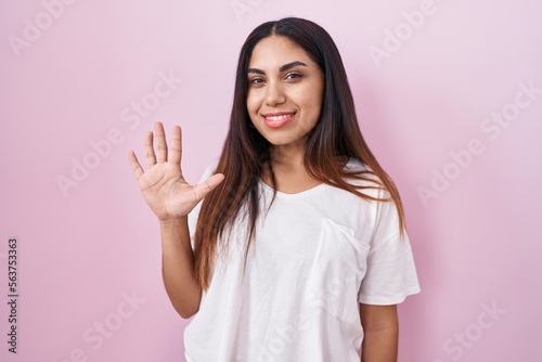 Young arab woman standing over pink background showing and pointing up with fingers number five while smiling confident and happy.