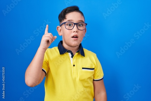 Young hispanic kid standing over blue background pointing finger up with successful idea. exited and happy. number one.