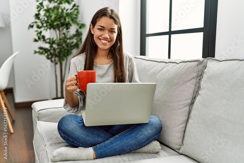 Young latin woman using laptop and drinking coffee sitting on sofa at home