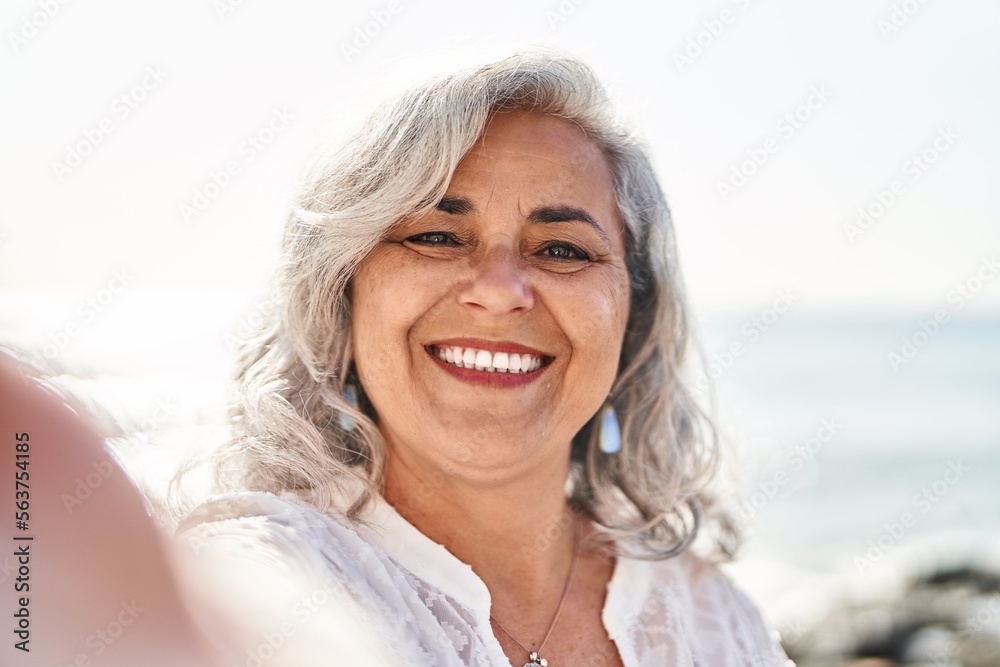 Middle age woman smiling confident making selfie by the camera at seaside