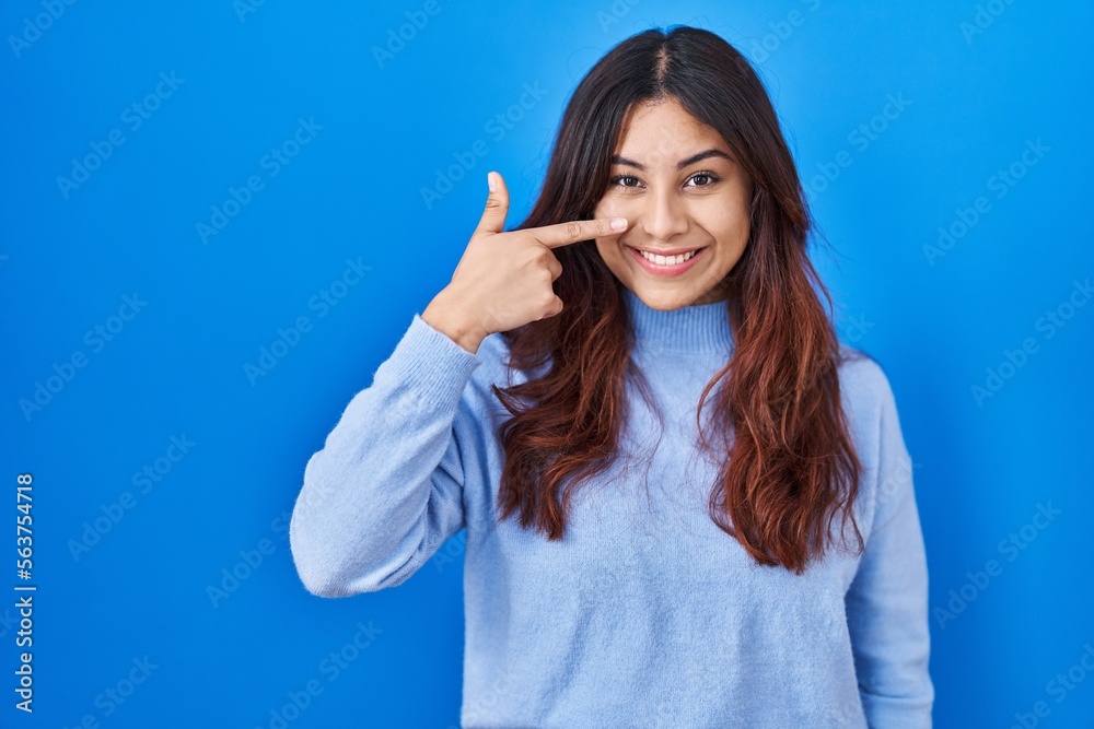 Hispanic young woman standing over blue background pointing with hand finger to face and nose, smiling cheerful. beauty concept Hispanic young woman standing over blue background pointing with hand finger to face and nose, smiling cheerful. beauty concept