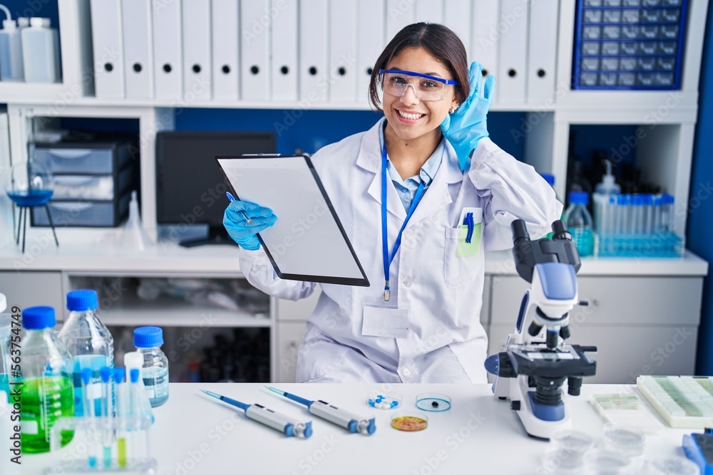 Hispanic young woman working at scientist laboratory smiling with hand over ear listening an hearing to rumor or gossip. deafness concept.