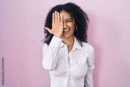 Hispanic woman with curly hair standing over pink background covering one eye with hand, confident smile on face and surprise emotion. © Krakenimages.com