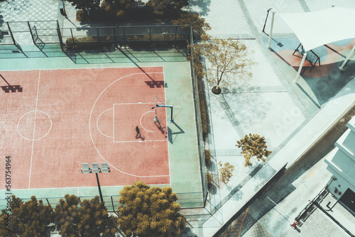 Aerial top view of a football/soccer and basketball fenced field, bordered by several trees, with two people training there, on a local school around Dubai photo