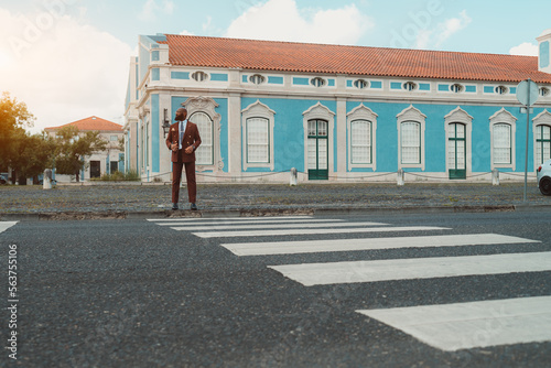A bald black man looking dapper in his Bordeaux custom-made suit standing in front of the zebrawalk waiting for the green light to cross the road, in front of a classical blue edifice