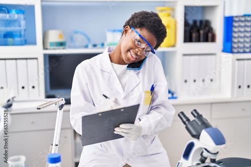 African american woman wearing scientist uniform talking on the smartphone write on clipboard at laboratory