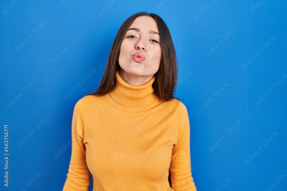 Young brunette woman standing over blue background looking at the camera blowing a kiss on air being lovely and sexy. love expression. Young brunette woman standing over blue background looking at the camera blowing a kiss on air being lovely and sexy. love expression.