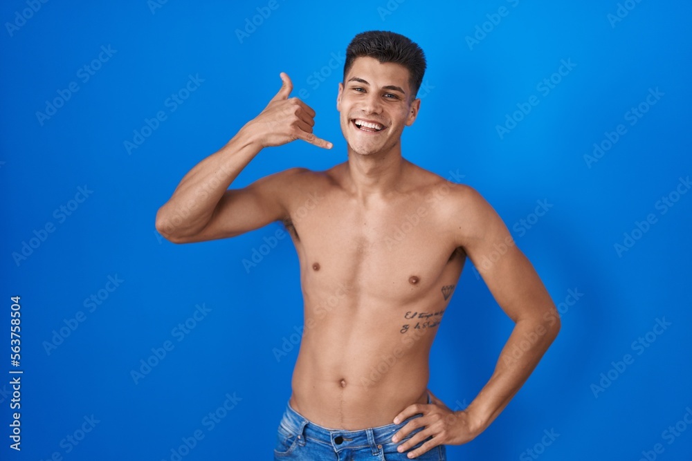 Young hispanic man standing shirtless over blue background smiling doing phone gesture with hand and fingers like talking on the telephone. communicating concepts. Young hispanic man standing shirtless over blue background smiling doing phone gesture with hand and fingers like talking on the telephone. communicating concepts.