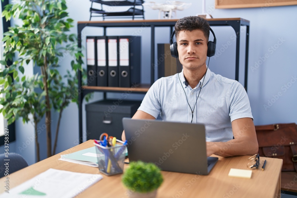 Young hispanic man working at the office wearing headphones relaxed with serious expression on face. simple and natural looking at the camera. Young hispanic man working at the office wearing headphones relaxed with serious expression on face. simple and natural looking at the camera.
