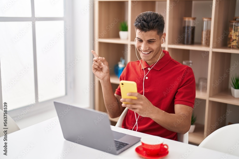 Young hispanic man using laptop and doing video call with smartphone smiling happy pointing with hand and finger to the side Young hispanic man using laptop and doing video call with smartphone smiling happy pointing with hand and finger to the side