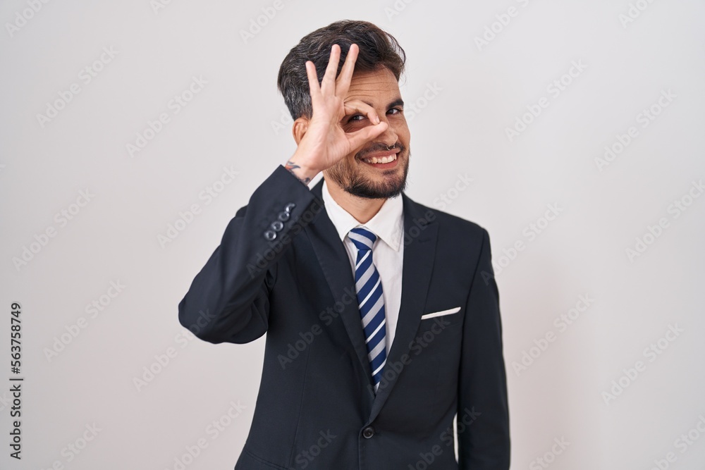 Young hispanic man with tattoos wearing business suit and tie doing ok gesture with hand smiling, eye looking through fingers with happy face. Young hispanic man with tattoos wearing business suit and tie doing ok gesture with hand smiling, eye looking through fingers with happy face.