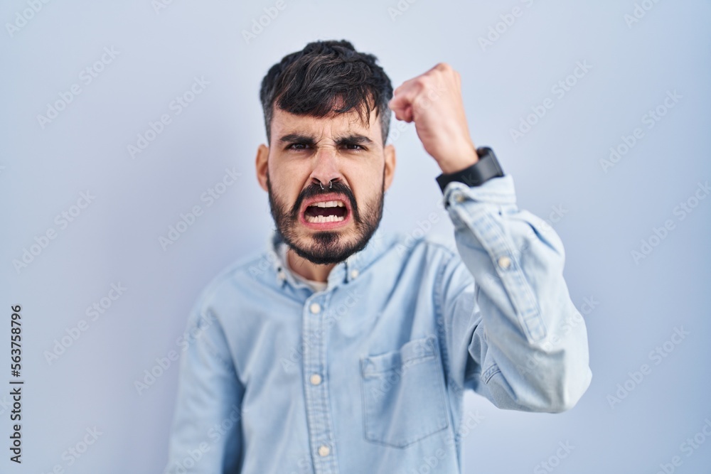 Young hispanic man with beard standing over blue background angry and mad raising fist frustrated and furious while shouting with anger. rage and aggressive concept. Young hispanic man with beard standing over blue background angry and mad raising fist frustrated and furious while shouting with anger. rage and aggressive concept.