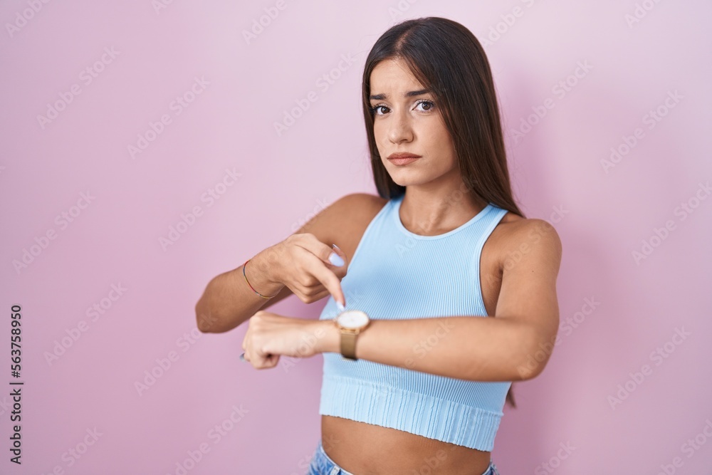 Young brunette woman standing over pink background in hurry pointing to watch time, impatience, upset and angry for deadline delay