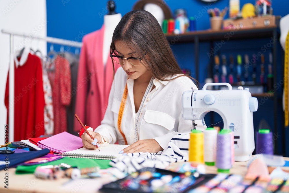 Fototapeta premium Young beautiful hispanic woman tailor drawing on notebook at clothing factory