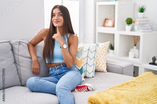 Young brunette woman sitting on the sofa at home thinking concentrated about doubt with finger on chin and looking up wondering