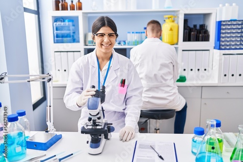 Young woman working at scientist laboratory looking positive and happy standing and smiling with a confident smile showing teeth