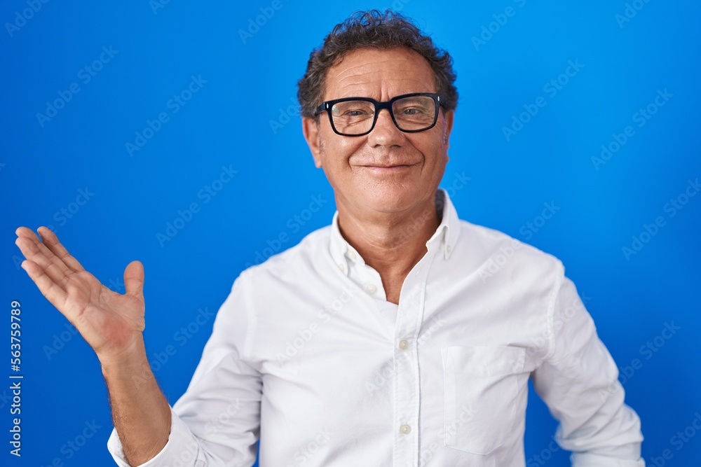 Middle age hispanic man standing over blue background smiling cheerful presenting and pointing with palm of hand looking at the camera. Middle age hispanic man standing over blue background smiling cheerful presenting and pointing with palm of hand looking at the camera.