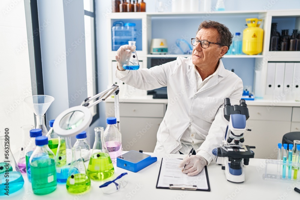 Middle age man wearing scientist uniform looking test tube writing on clipboard at laboratory Middle age man wearing scientist uniform looking test tube writing on clipboard at laboratory