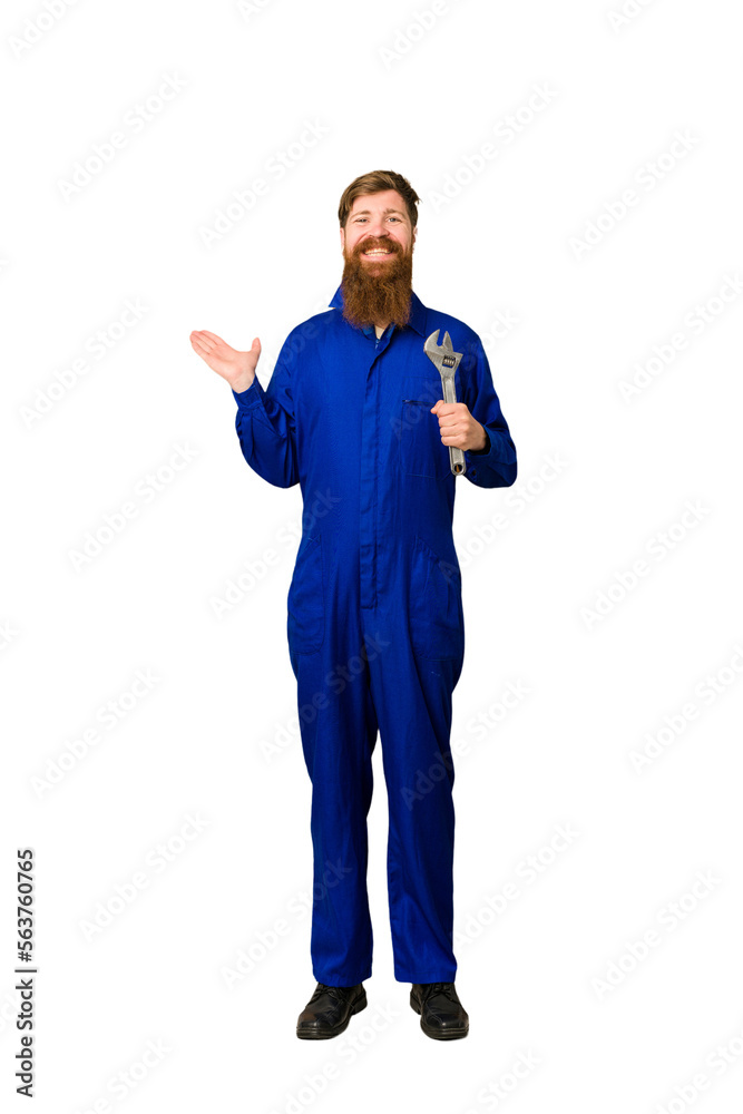 Young adult redhead mechanic man full body isolated showing a copy space on a palm and holding another hand on waist. Young adult redhead mechanic man full body isolated showing a copy space on a palm and holding another hand on waist.