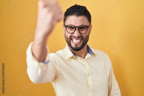 Hispanic young man wearing business clothes and glasses angry and mad raising fist frustrated and furious while shouting with anger. rage and aggressive concept.
