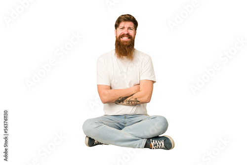 Young adult redhead with a long beard sitting on the floor isolated who feels confident, crossing arms with determination.