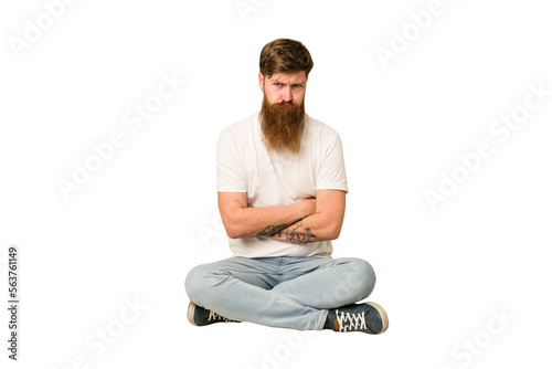 Young adult redhead with a long beard sitting on the floor isolated unhappy looking in camera with sarcastic expression.