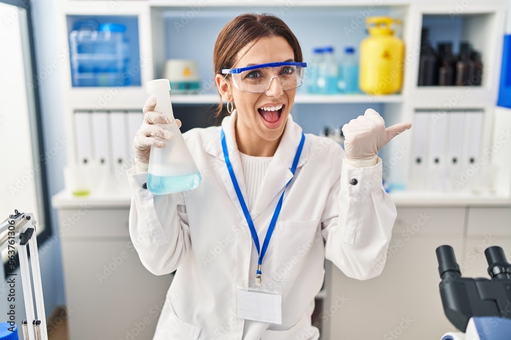 Young brunette woman working at scientist laboratory pointing thumb up to the side smiling happy with open mouth Young brunette woman working at scientist laboratory pointing thumb up to the side smiling happy with open mouth