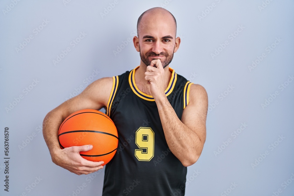 Young bald man with beard wearing basketball uniform holding ball looking confident at the camera smiling with crossed arms and hand raised on chin. thinking positive.