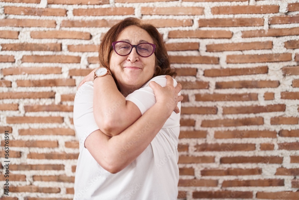 Senior woman with glasses standing over bricks wall hugging oneself happy and positive, smiling confident. self love and self care Senior woman with glasses standing over bricks wall hugging oneself happy and positive, smiling confident. self love and self care