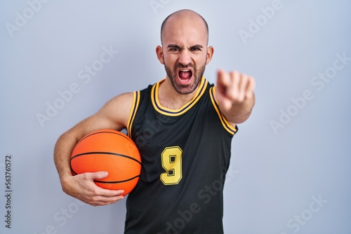 Young bald man with beard wearing basketball uniform holding ball pointing displeased and frustrated to the camera, angry and furious with you