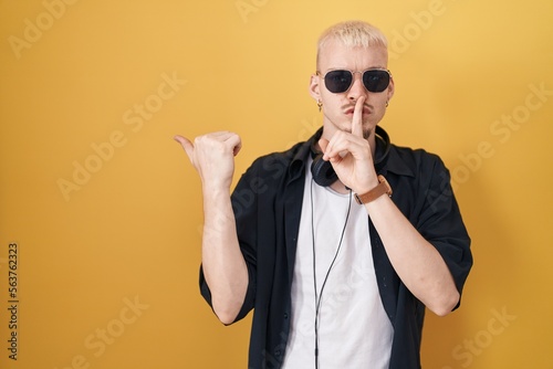 Young caucasian man wearing sunglasses standing over yellow background asking to be quiet with finger on lips pointing with hand to the side. silence and secret concept. © Krakenimages.com