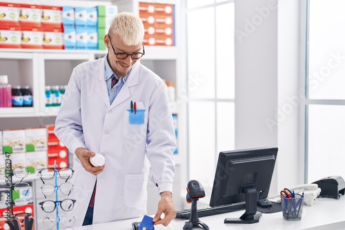 Young caucasian man pharmacist holding pills bottle using data phone and credit card at pharmacy