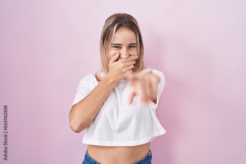 Young blonde woman standing over pink background laughing at you, pointing finger to the camera with hand over mouth, shame expression Young blonde woman standing over pink background laughing at you, pointing finger to the camera with hand over mouth, shame expression