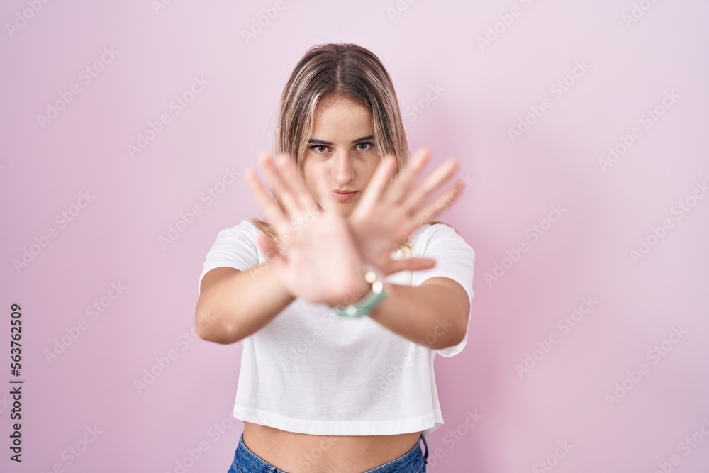 Young blonde woman standing over pink background rejection expression crossing arms and palms doing negative sign, angry face