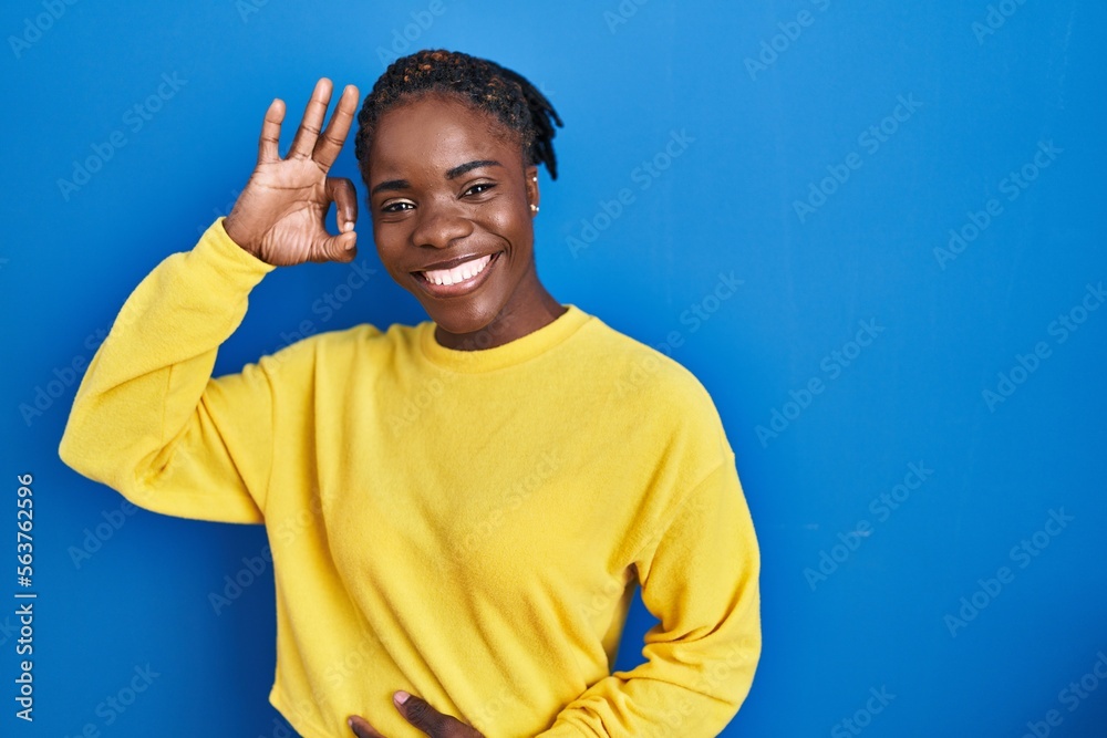 Naklejka premium Beautiful black woman standing over blue background smiling positive doing ok sign with hand and fingers. successful expression.