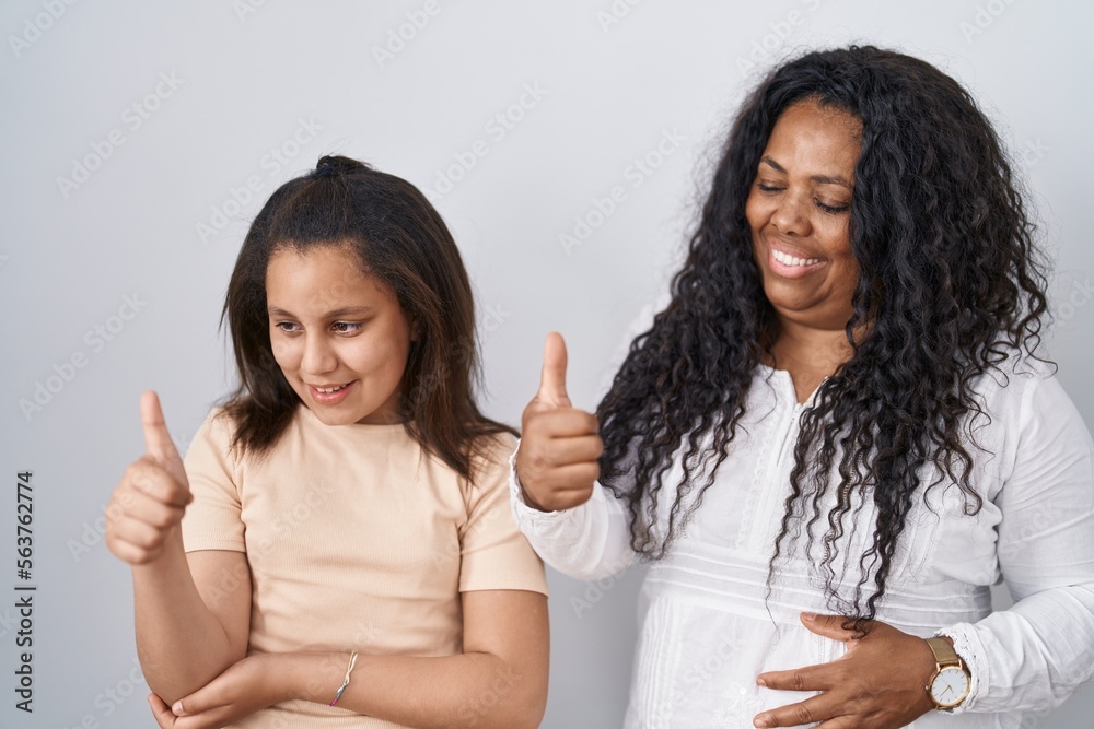 Mother and young daughter standing over white background looking proud, smiling doing thumbs up gesture to the side Mother and young daughter standing over white background looking proud, smiling doing thumbs up gesture to the side