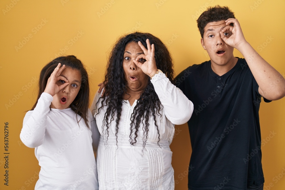 Family of mother, daughter and son standing over yellow background doing ok gesture shocked with surprised face, eye looking through fingers. unbelieving expression. Family of mother, daughter and son standing over yellow background doing ok gesture shocked with surprised face, eye looking through fingers. unbelieving expression.