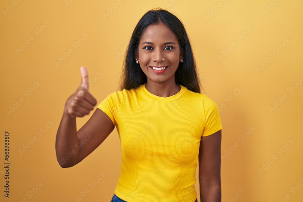 Young indian woman standing over yellow background doing happy thumbs up gesture with hand. approving expression looking at the camera showing success. Young indian woman standing over yellow background doing happy thumbs up gesture with hand. approving expression looking at the camera showing success.