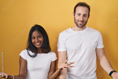 Interracial couple standing over yellow background smiling cheerful with open arms as friendly welcome, positive and confident greetings