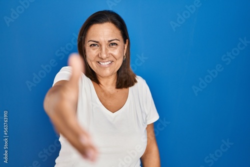 Hispanic mature woman standing over blue background smiling friendly offering handshake as greeting and welcoming. successful business. © Krakenimages.com