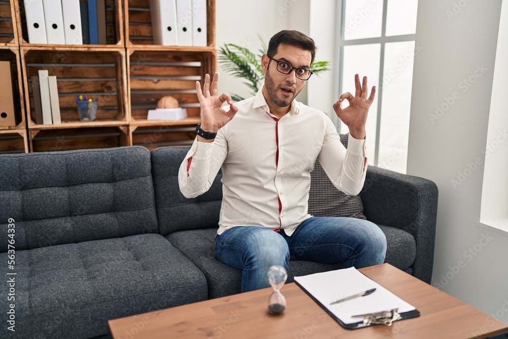 Young hispanic man with beard working at consultation office looking surprised and shocked doing ok approval symbol with fingers. crazy expression Young hispanic man with beard working at consultation office looking surprised and shocked doing ok approval symbol with fingers. crazy expression